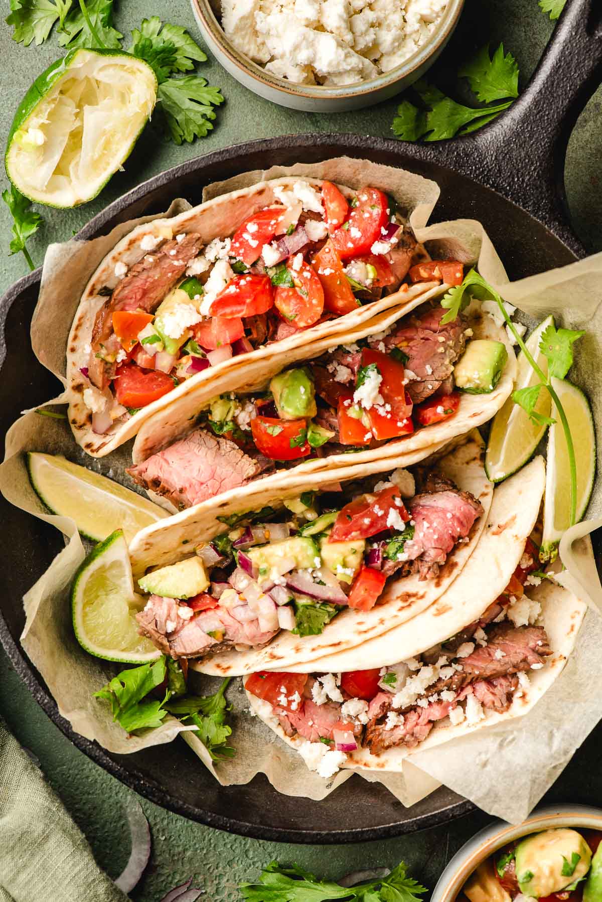 Four flank steak tacos nestled in a cast iron skillet surrounded with lime wedges.