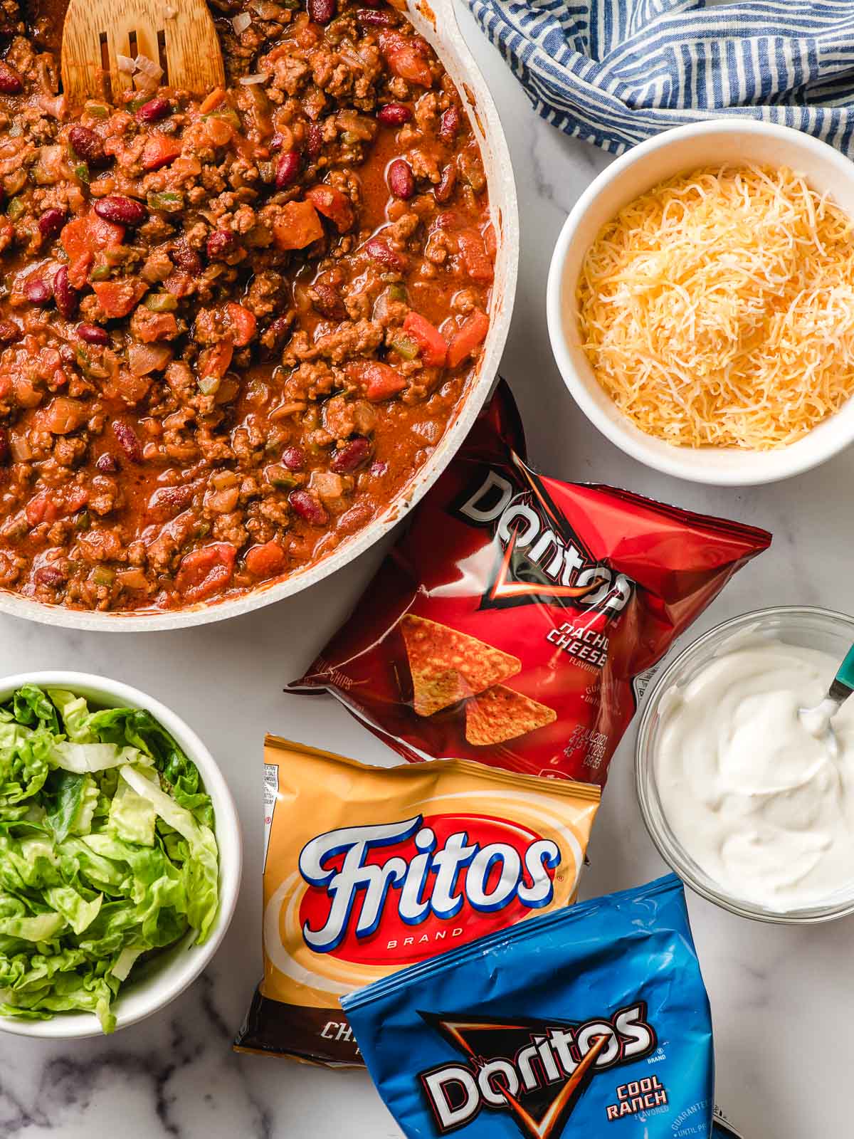 Meat mixture for Walking Tacos with bags of Doritos and Fritos, plus bowls of lettuce, cheese, and sour cream on the side.