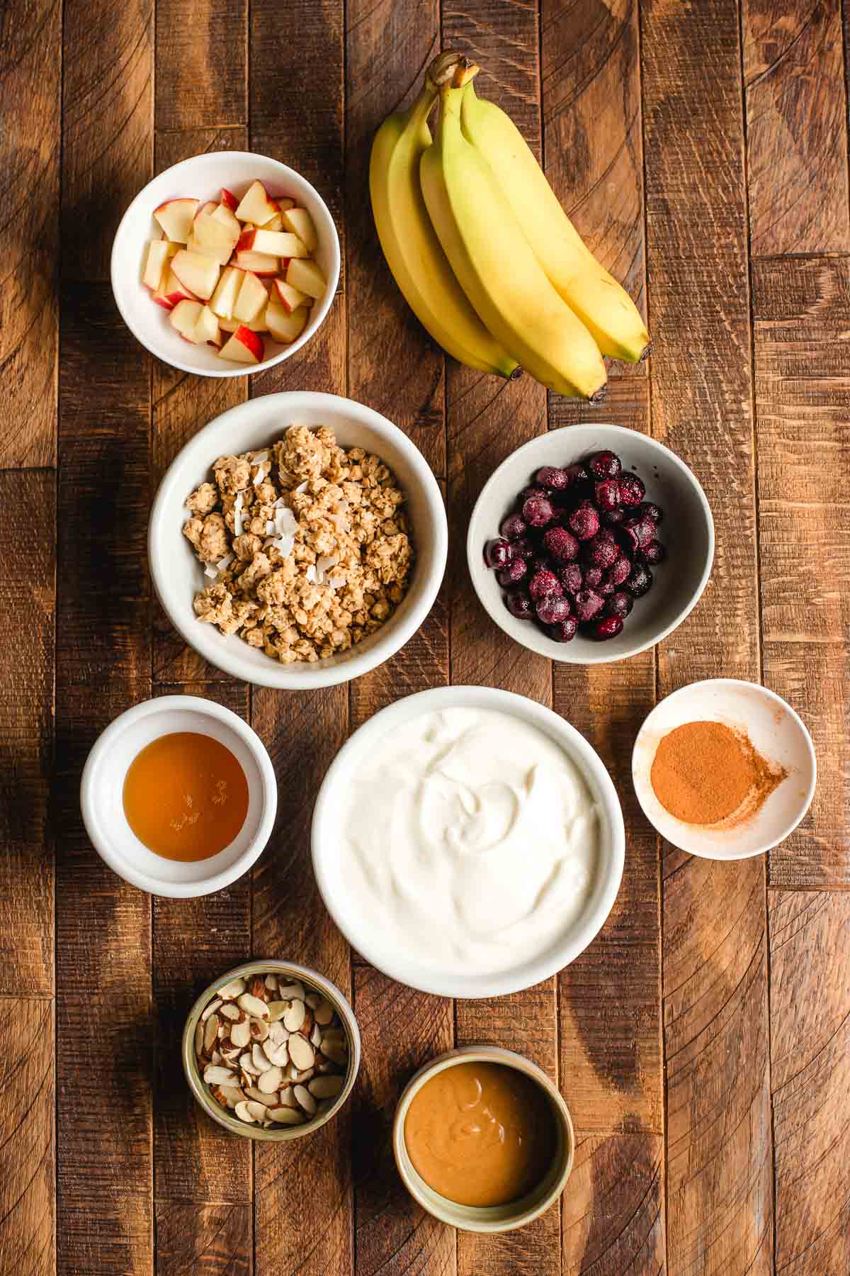 Bowls of yogurt, bananas, blueberries, apples, cinnamon, almonds, granola, and honey serried on a wooden board.