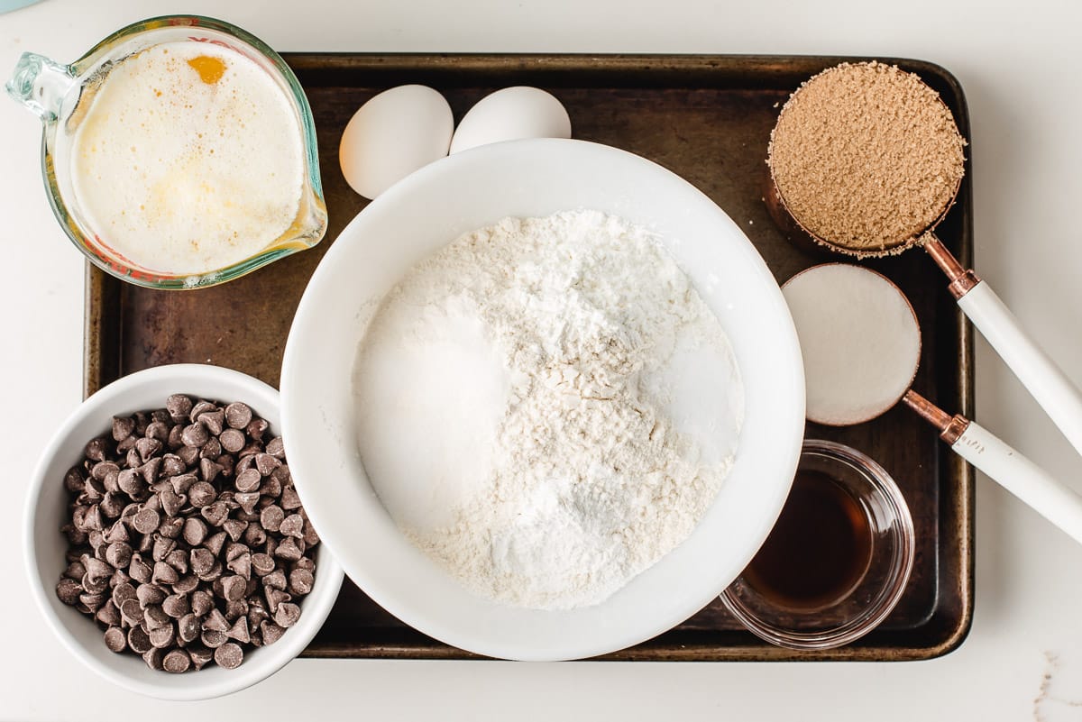 Bowls of flour, baking soda, salt, cornstarch, brown sugar, sugar, melted butter, eggs, vanilla, and chocolate chips.