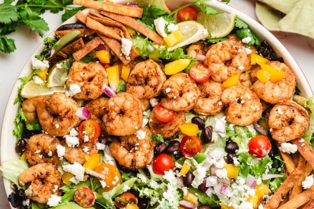Seasoned shrimp, tortilla strips, lime, black beans, queso fresco, and mixed greens in a bowl.