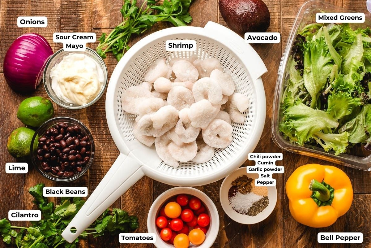 Shrimp, bell pepper, tomatoes, corn, avocado, greens, red onion, black beans, cilantro, and spices in bowls on a wood background.