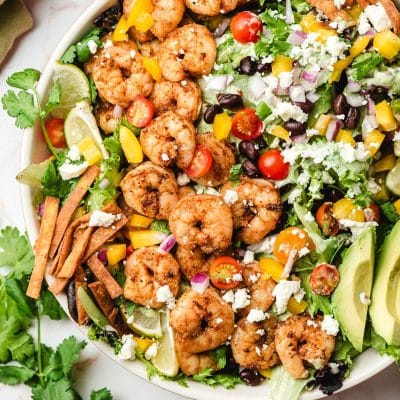 Mixed greens topped with shrimp, tortilla strips, avocado, cheese, and fresh veggies in a white bowl.