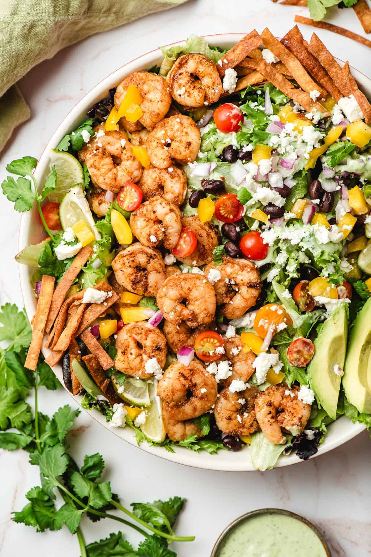 Mixed greens topped with shrimp, tortilla strips, avocado, cheese, and fresh veggies in a white bowl.