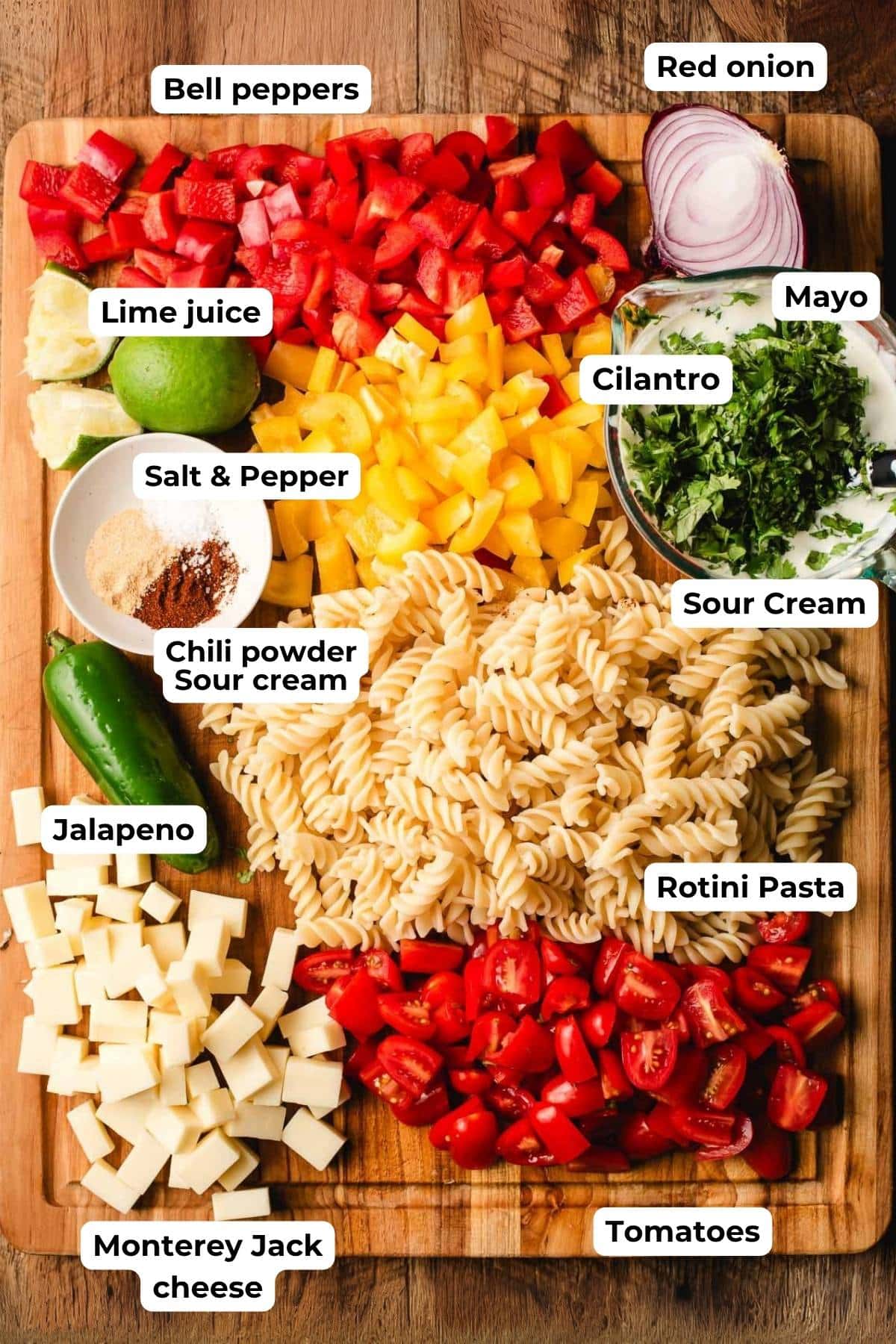 Bell peppers, red onion, limes, cooked pasta, cheese cubes, spices, tomatoes, and a creamy cilantro dressing on a cutting board.