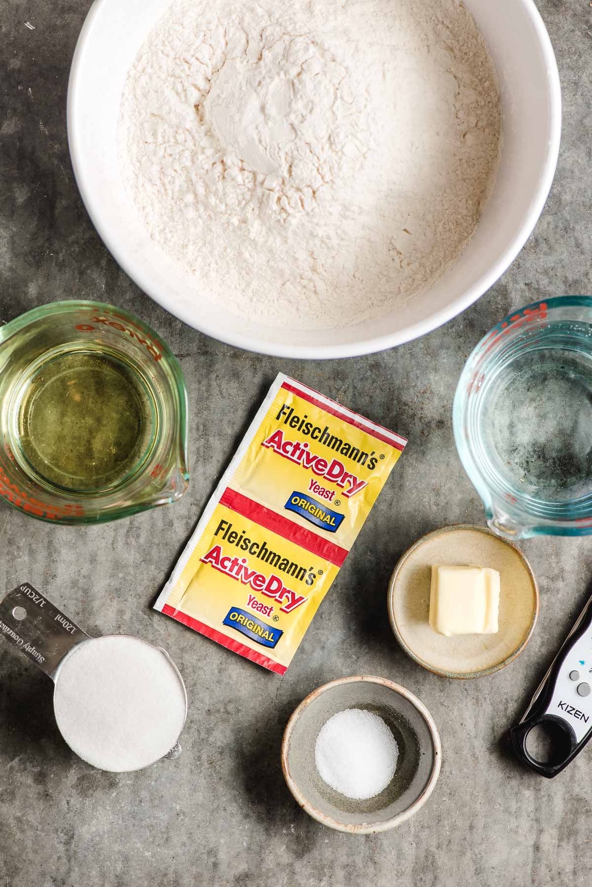 Baking ingredients displayed in small bowls- flour, vegetable oil, yeast, salt, sugar, water, and butter.