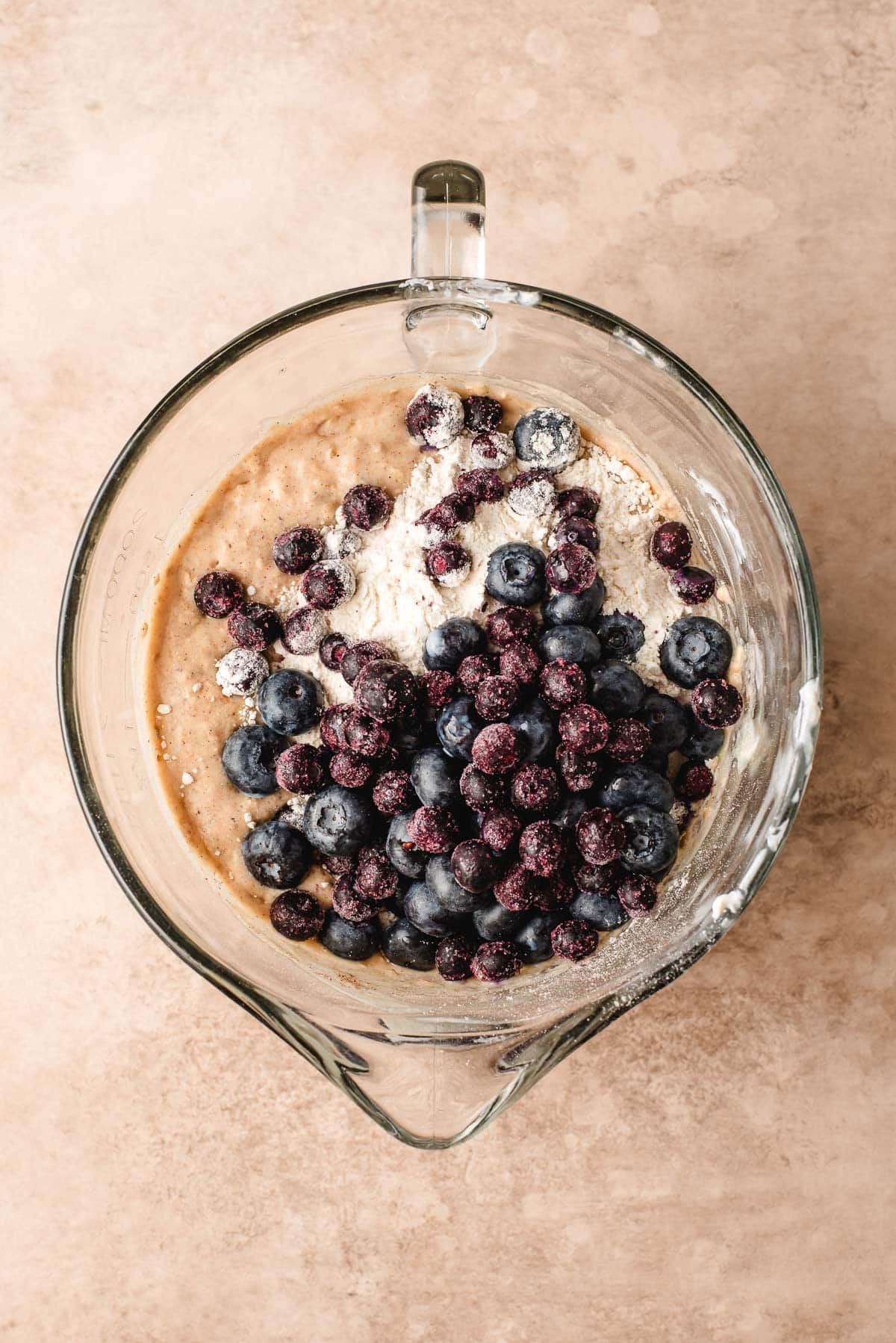 Blueberries and flour poured on top of muffin batter in a glass mixing bowl.