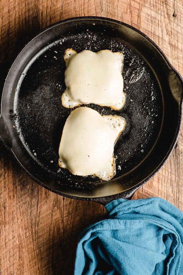 Cast iron skillet with two burgers covered in melted Muenster cheese.