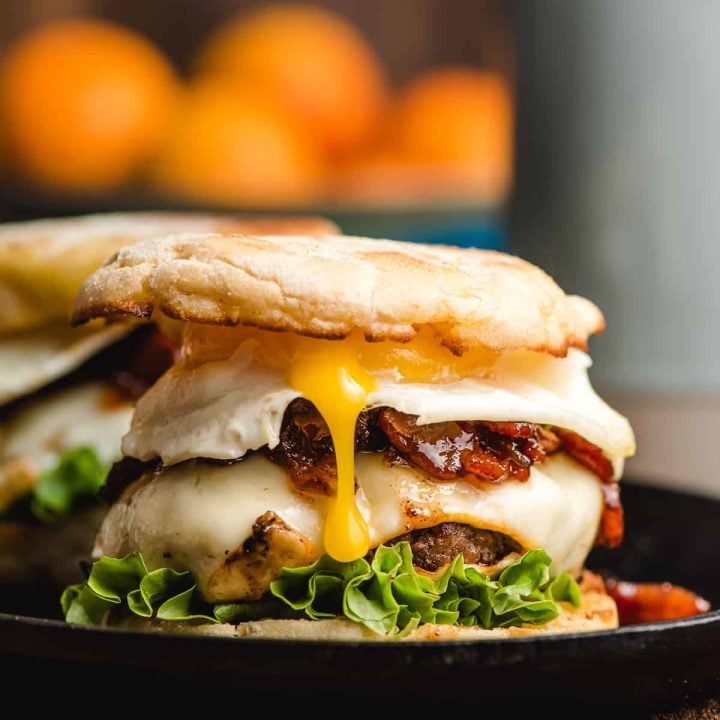 Breakfast burger featuring an English muffin, cheeseburger, bacon jam, and a fried egg with the yolk spilling out the side.