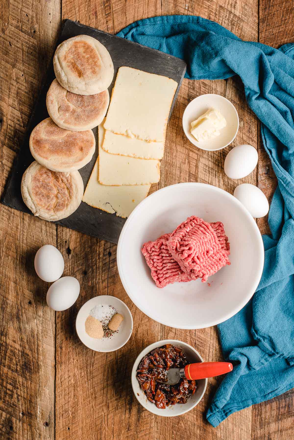 English muffins, cheese slices, ground beef, eggs, bacon jam, and seasonings shown on a wood background.