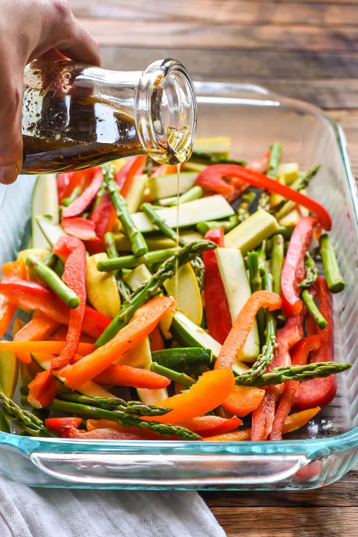 Vegetables in a glass baking dish with bottle pouring balsamic marinade on top, featured as a side for burgers and hot dogs.