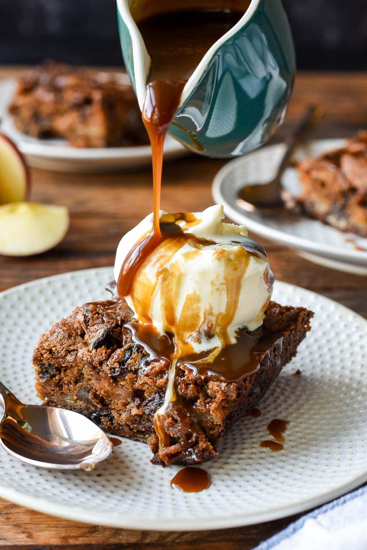 a piece of spiced apple cake on a plate, topped with ice cream and being drizzled with caramel sauce