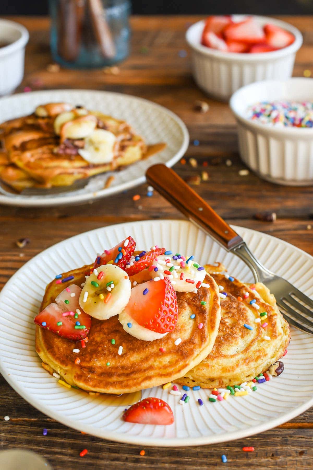 Two pancakes on a plate topped with sprinkles and banana and strawberry slices.