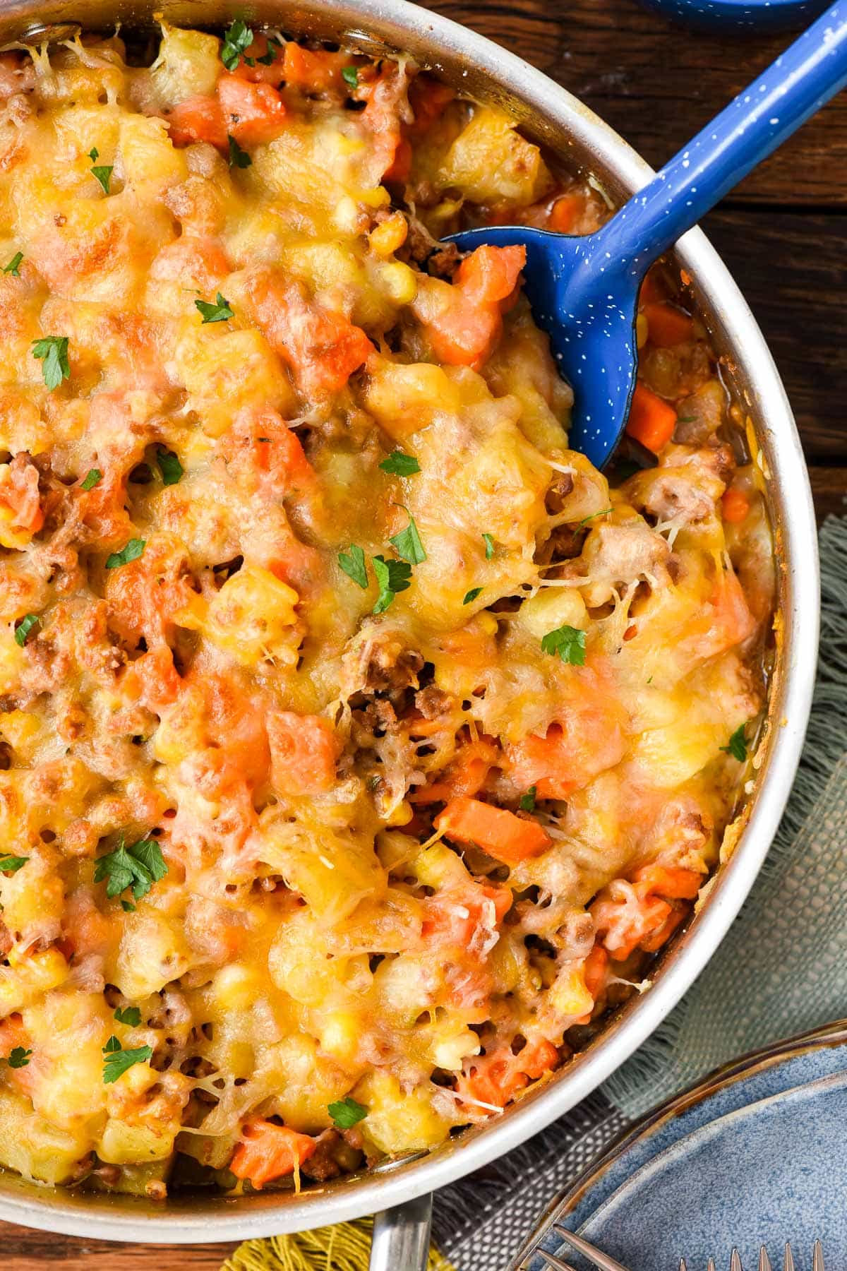Skillet of ground beef, potatoes, and corn topped with cheese with a blue spoon scooping into it.
