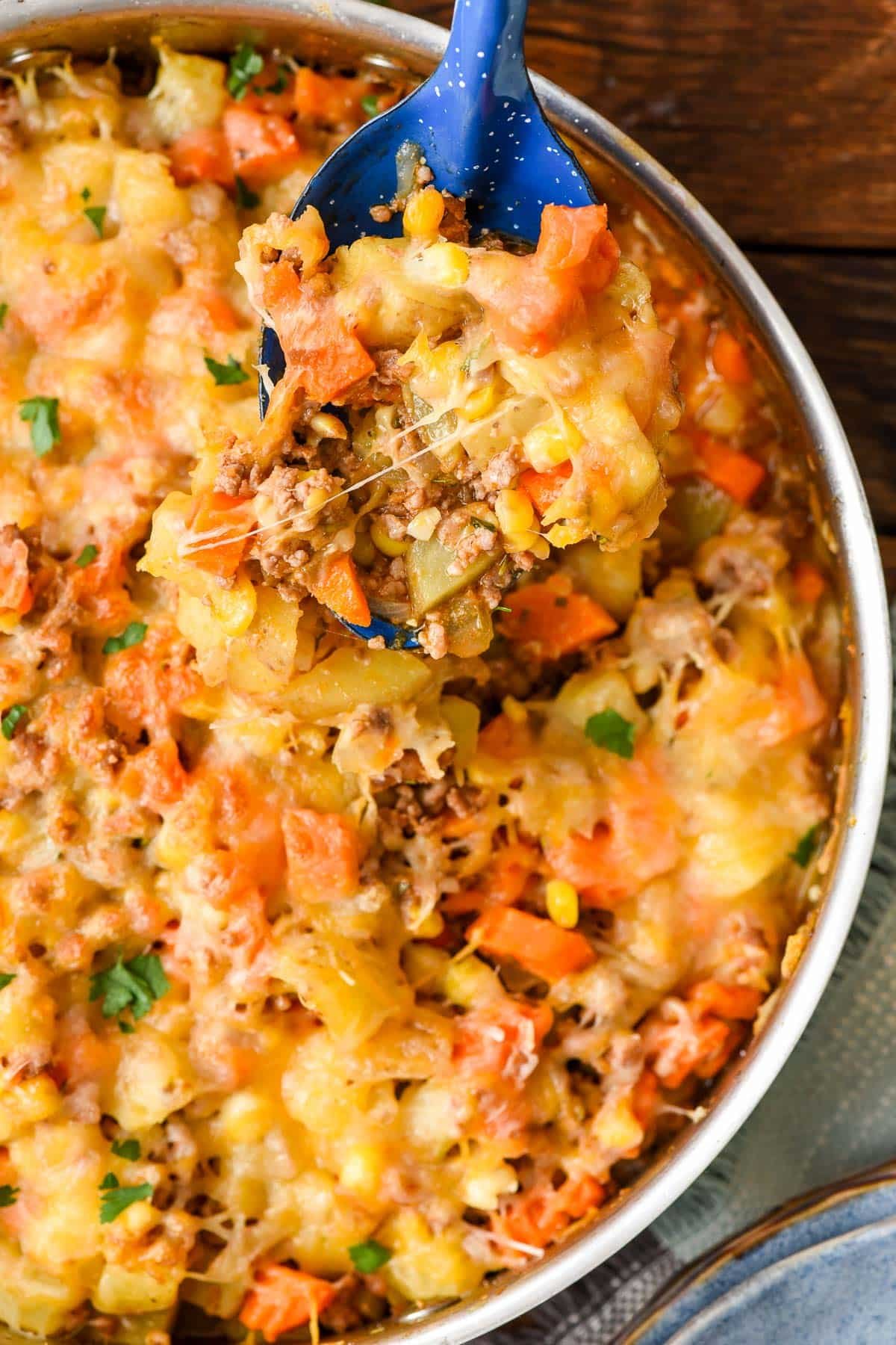 Blue spoon scooping cheesy ground beef and potatoes out of a skillet.