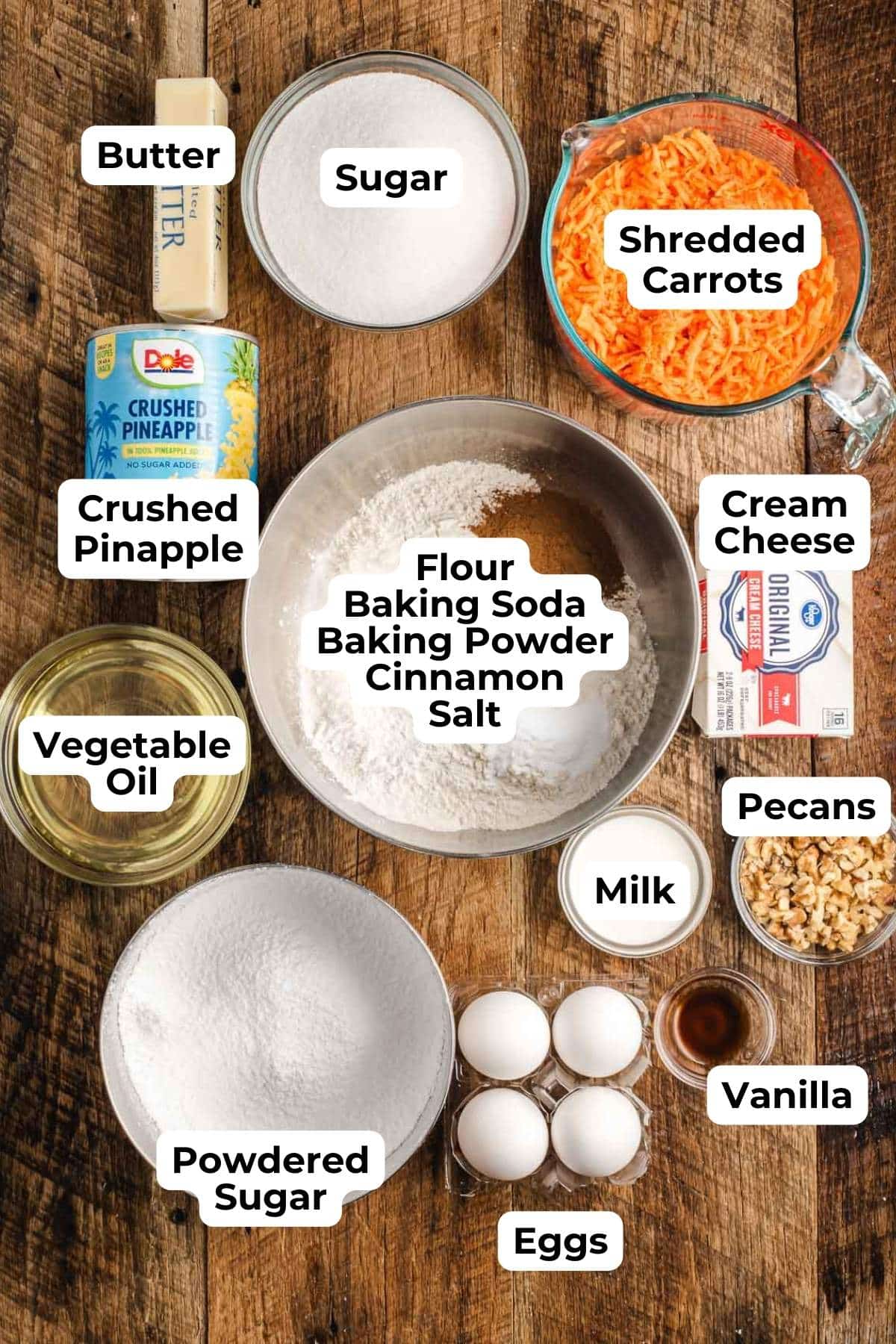 Butter, sugar, shredded carrots, canned pineapple, baking staples, eggs, milk, pecans, cream cheese, and vegetable oil in glass bowls on a wooden background.