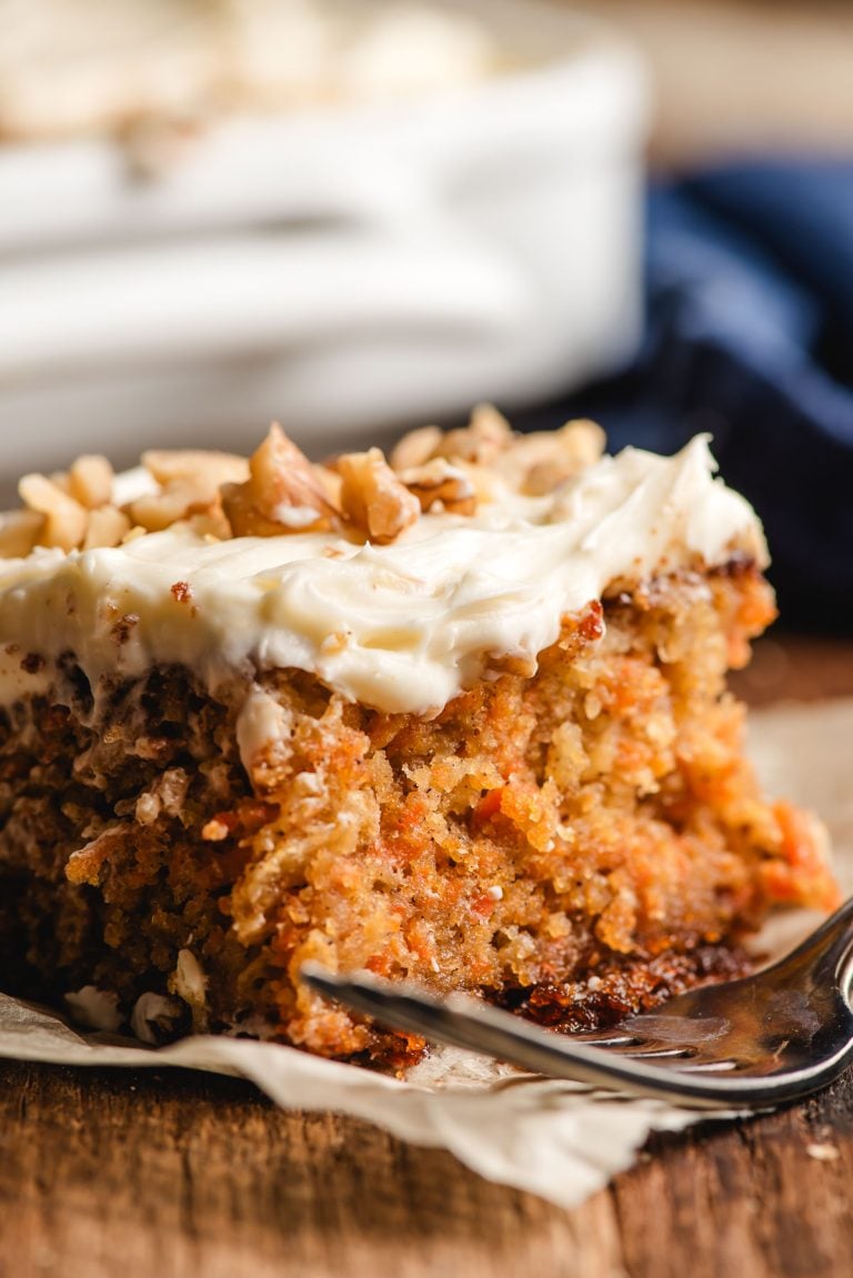 Slice of carrot sheet cake with cream cheese frosting, sprinkled with pecans.