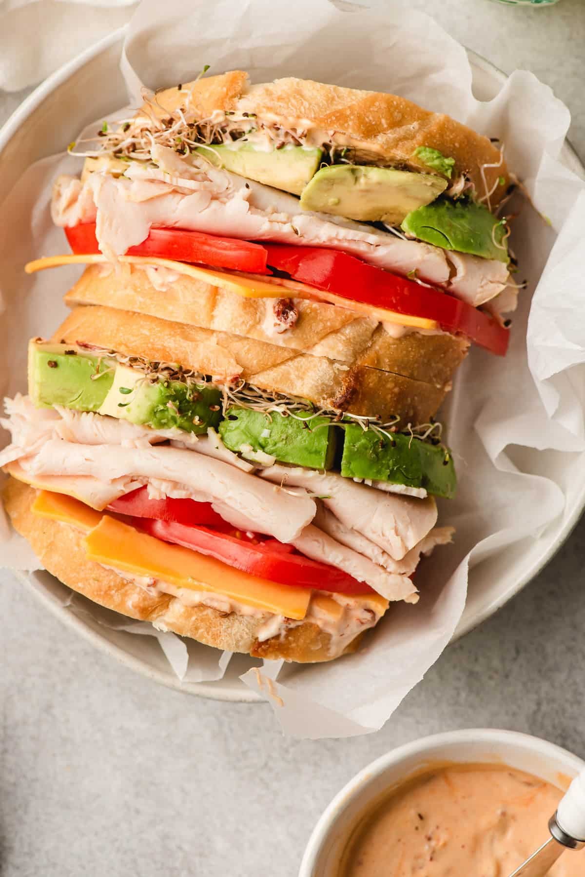 Two sandwiches with turkey, avocado, cheese, tomato, and sprouts in a bowl.