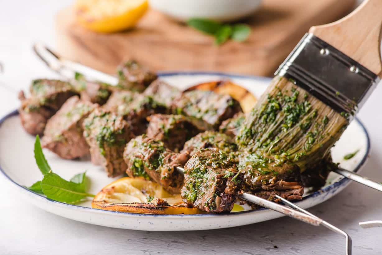 Grilled beef skewers being brushed with mint chimichurri