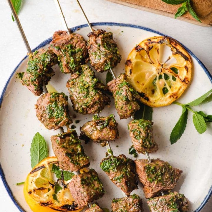 Steak kabobs on a plate with basil mint chimichuri and lemon.