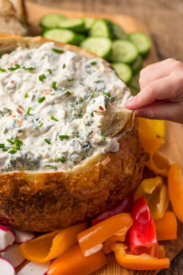 hand dipping a bread chunk into a bread bowl full of spinach dip surrounded by fresh sliced vegetables