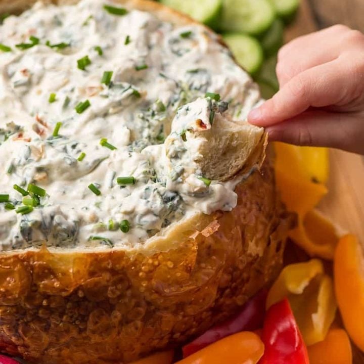 hand dipping a bread chunk into a bread bowl of spinach dip surrounded by fresh vegetables