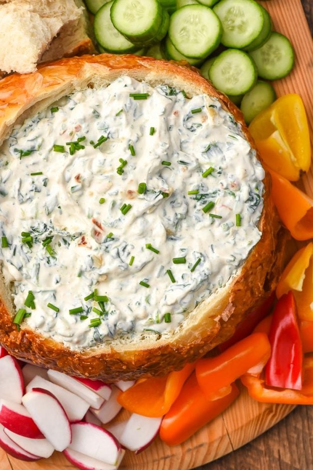 spinach dip inside a bread bowl surrounded by fresh sliced vegetables