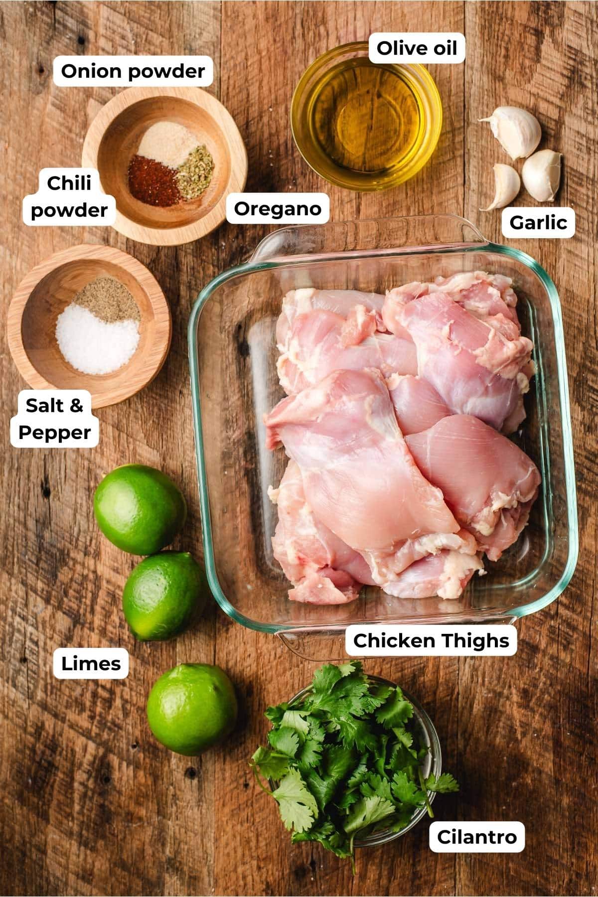 Bowls of chili powder, oregano, onion powder, salt and pepper. Limes, cilantro, olive oil, garlic cloves, and chicken thighs.