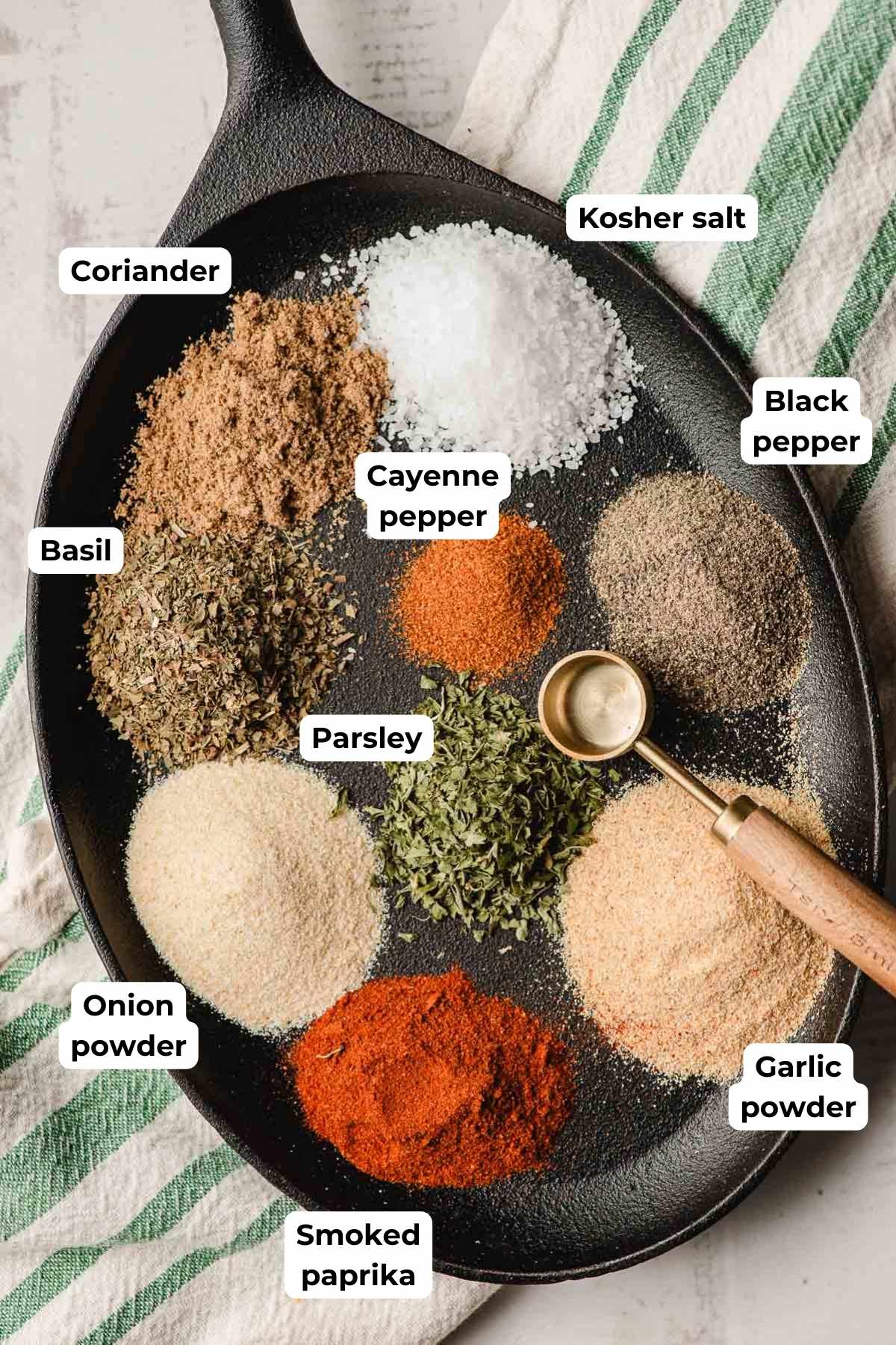 Kosher salt, pepper, garlic powder, smoked paprika, onion powder, dried basil, dried parsley, coriander, and cayenne pepper on a black cast iron platter.