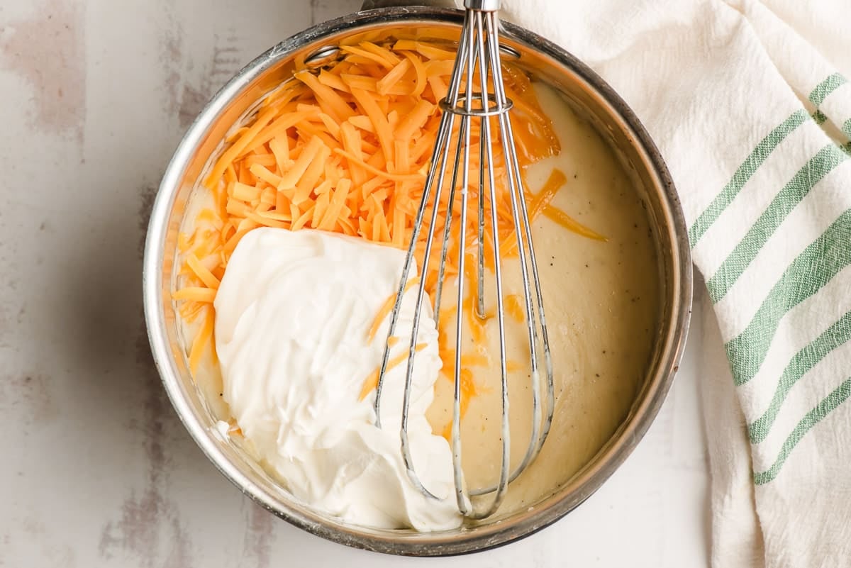 Shredded cheddar cheese and sour cream stirred into a creamy white sauce in a sauce pan.