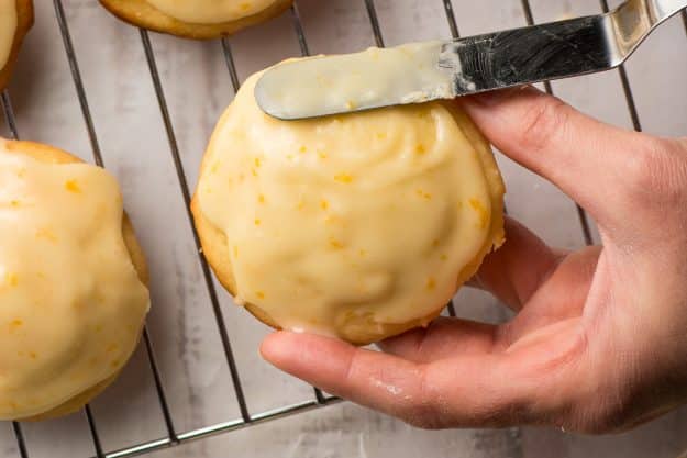 Hand holding an orange cookie and spreading glaze on it with an offset spatula.