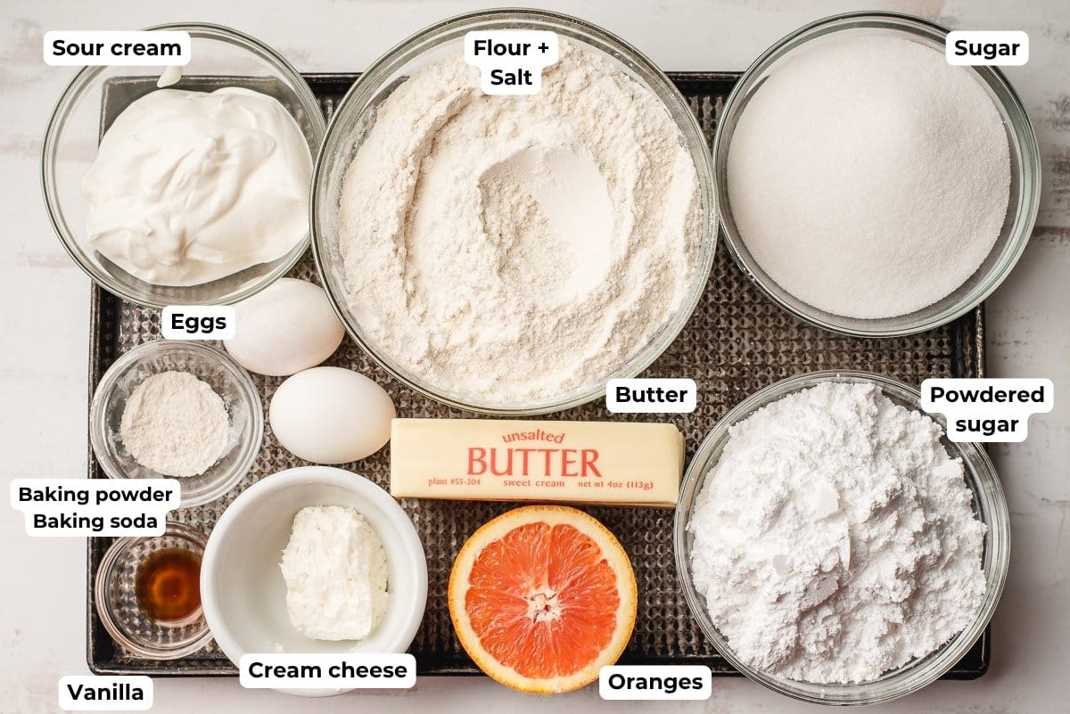 Sour cream, flour, sugar, baking powder and baking soda, eggs, butter, vanilla, cream cheese, orange half, and powdered sugar in glass bowls.