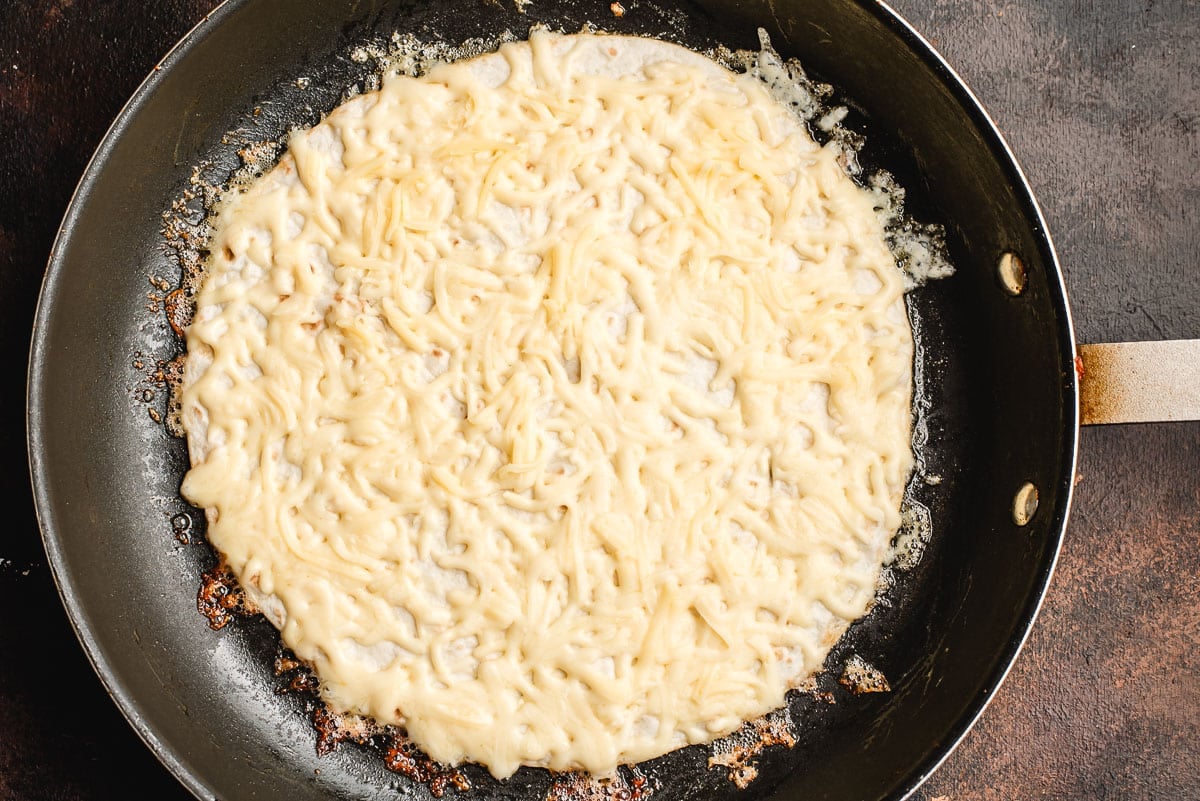 Shredded cheese melting on top of a flour tortilla in a pan.