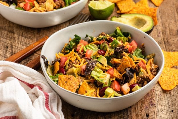 a bowl of doritos taco salad sits on a table with napkin and other ingredients