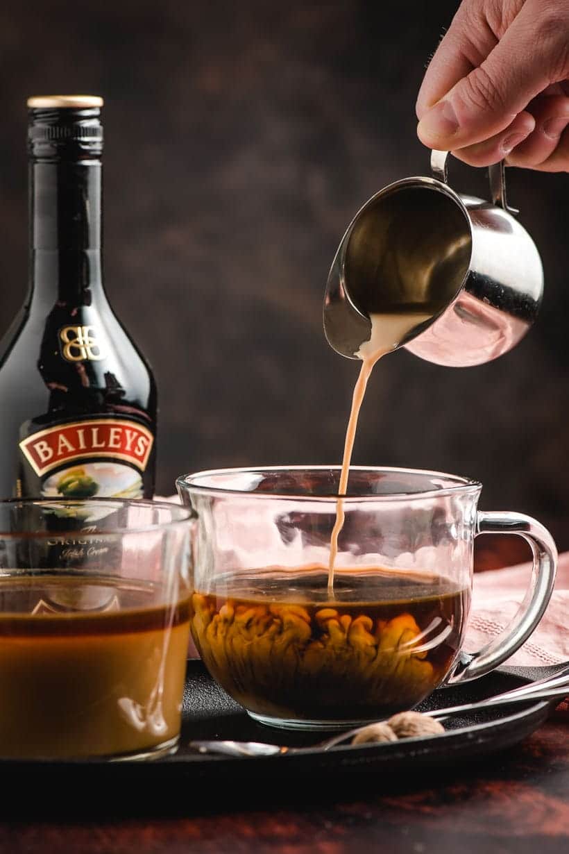 Metal pitcher pouring Bailey's Irish cream in coffee, with swirls of cream billowing up into the coffee.