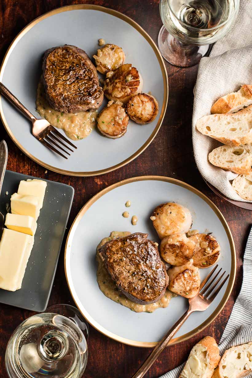 Two gold rimmed plates with steak and scallops, plus bread slices and butter on the side.