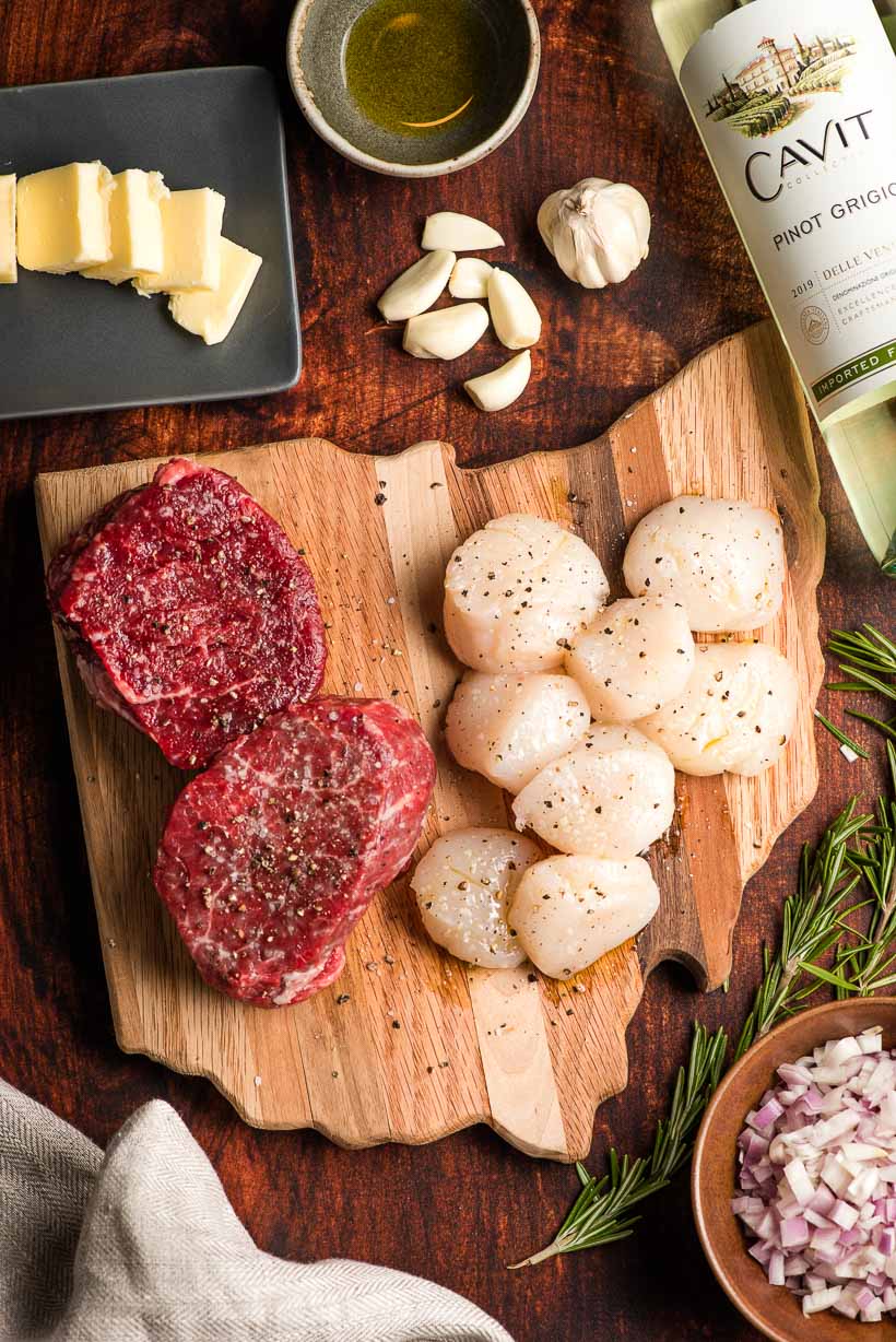 Ingredients for a surf and turf meal displayed on a cutting board- steak, scallops, butter, garlic, wine, shallots, and rosemary.