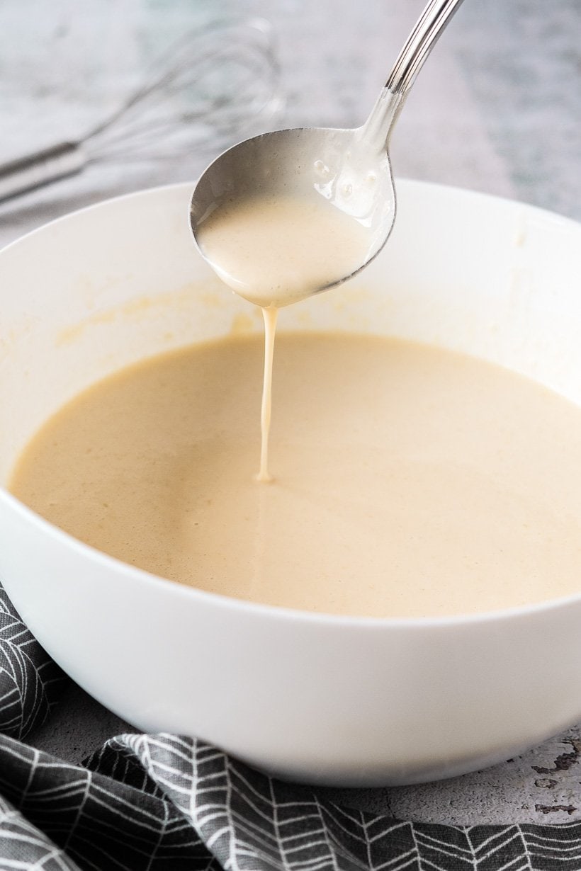Crepe batter in a white bowl with spoon scooping some batter.