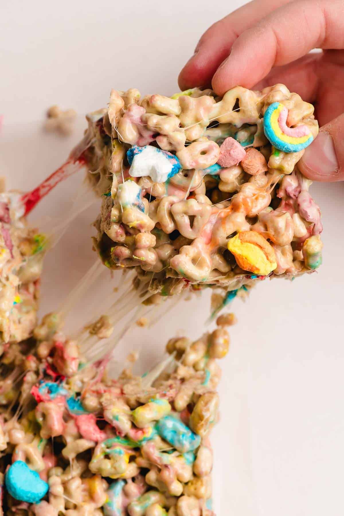 Hand pulling a Lucky Charms Krispies Treat away from the pan, leaving strings of gooey marshmallow behind.