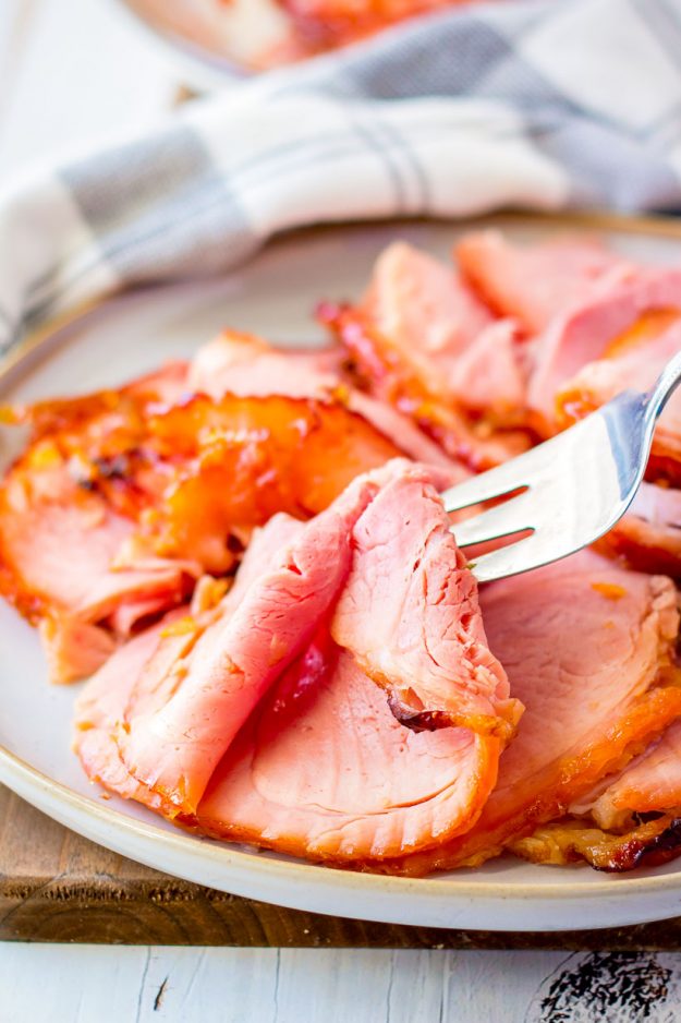 a fork lifts a slice of orange glazed ham off a serving plate