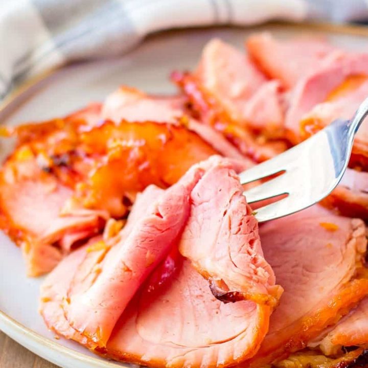 a fork lifts a slice of orange glazed ham off a serving plate