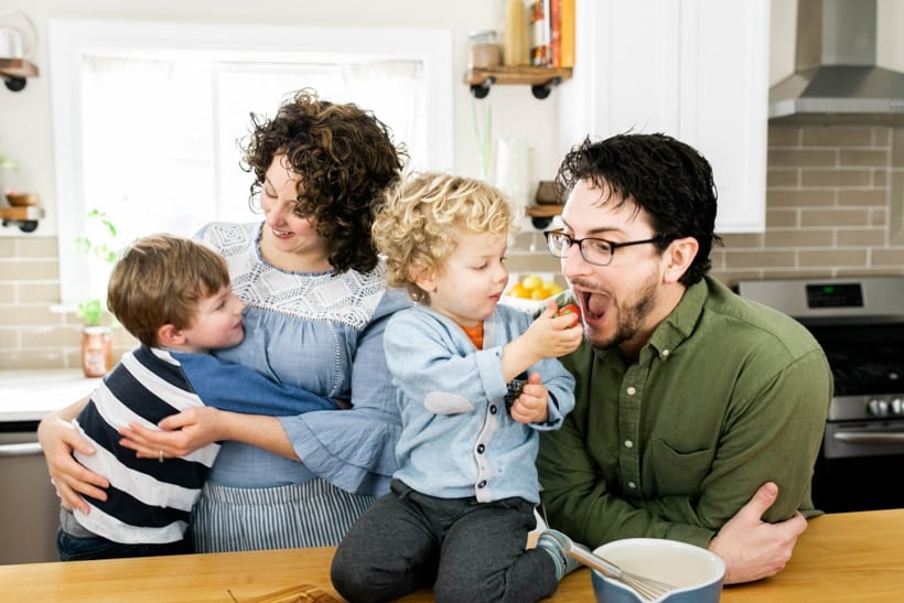 A young blonde boy hugging a white woman with curly dark hair. Another curly headed blonde toddler feedingn a strawberry to a white man with glasses.