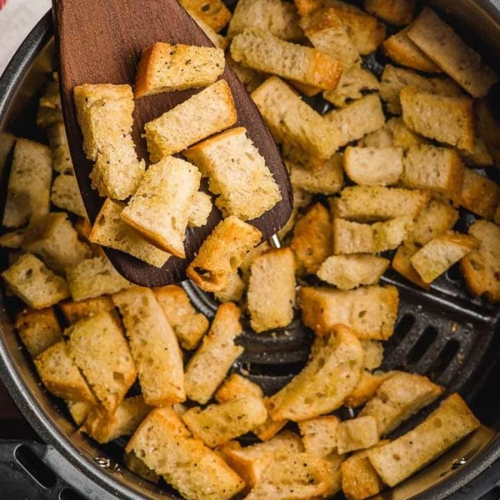 Wooden spatula scooping croutons out of an air fryer basket.