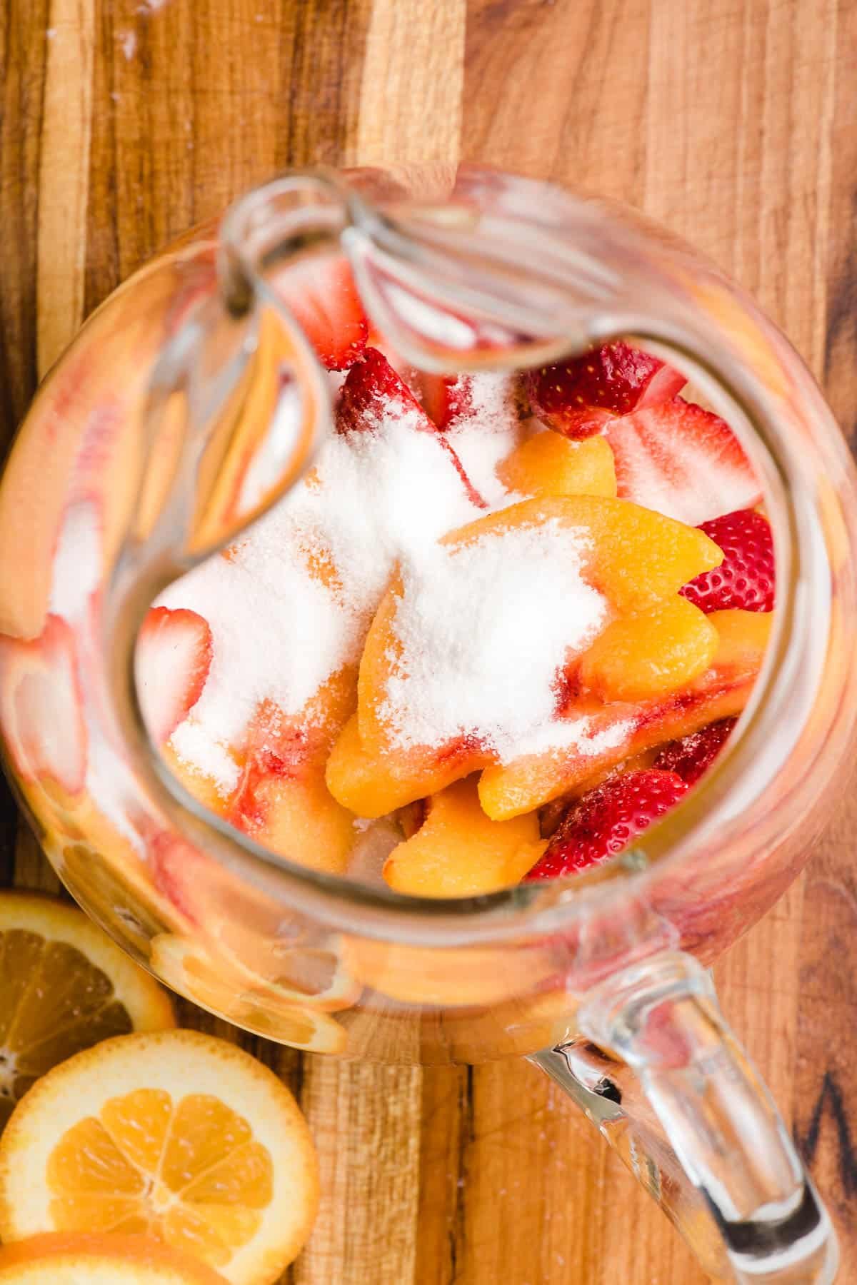 Sugar poured over strawberries and peaches in a glass pitcher.
