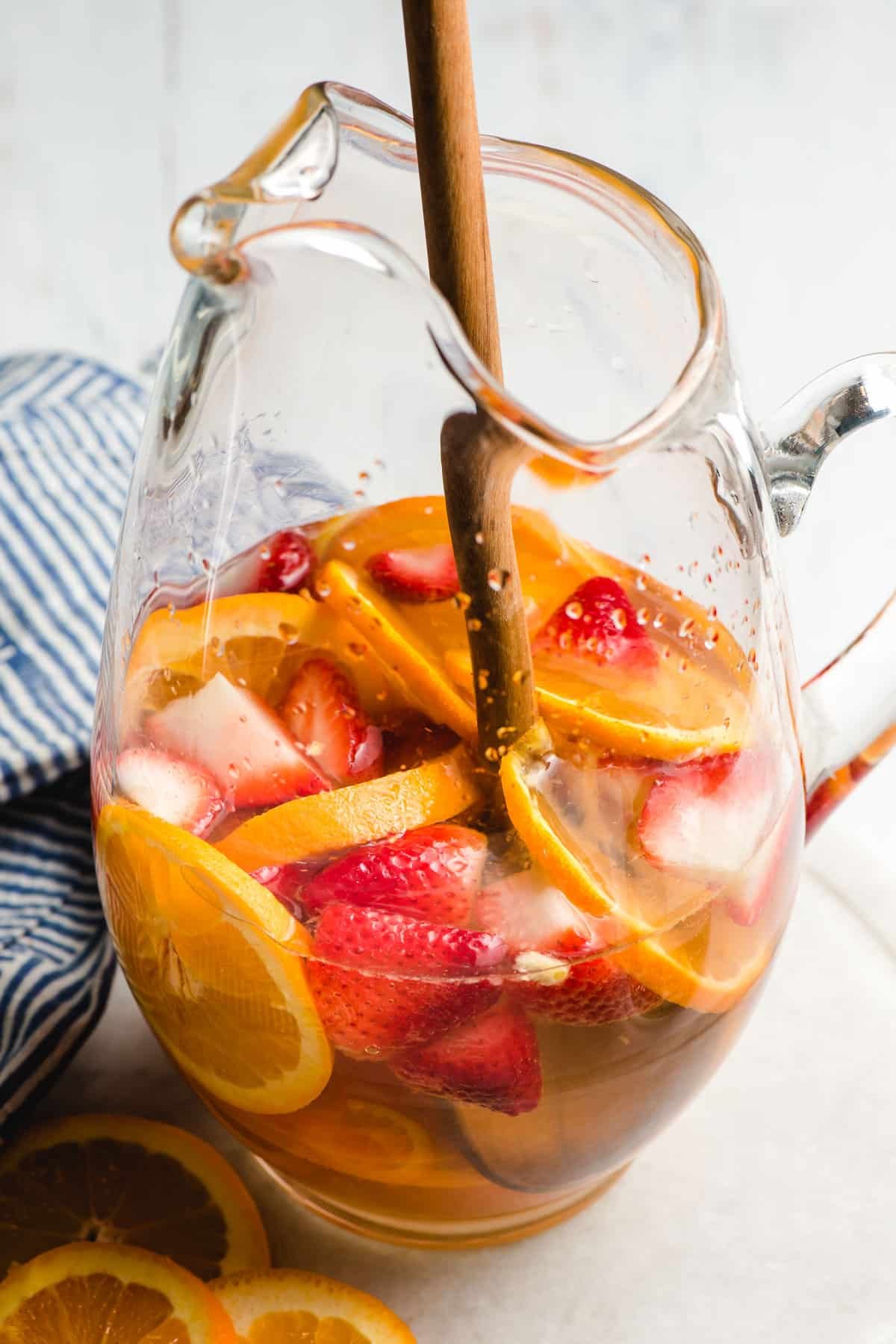 Wooden spoon stirring fresh fruits and wine together in a glass pitcher.