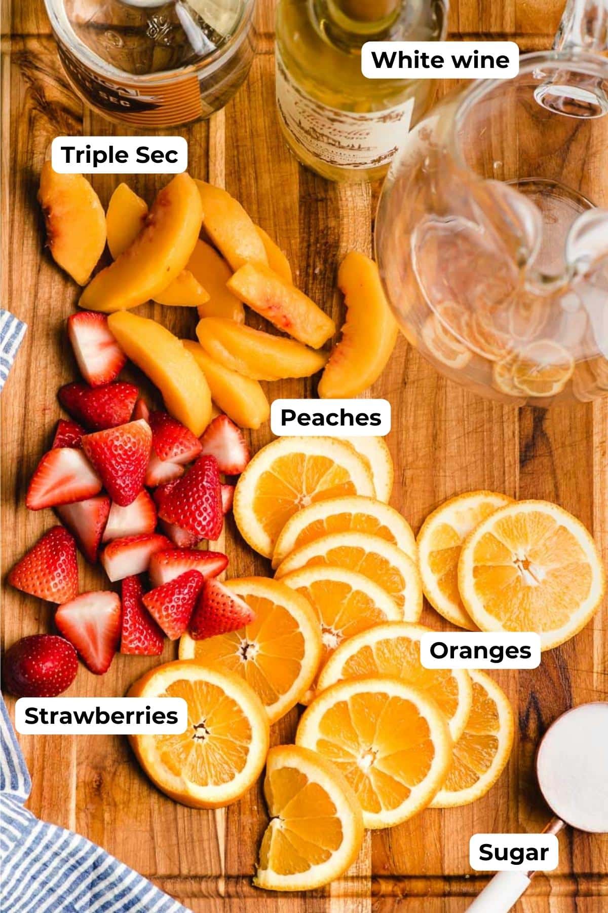Cutting board with a bottle of triple sec, white wine, sliced peaches, strawberries, and oranges, and sugar.