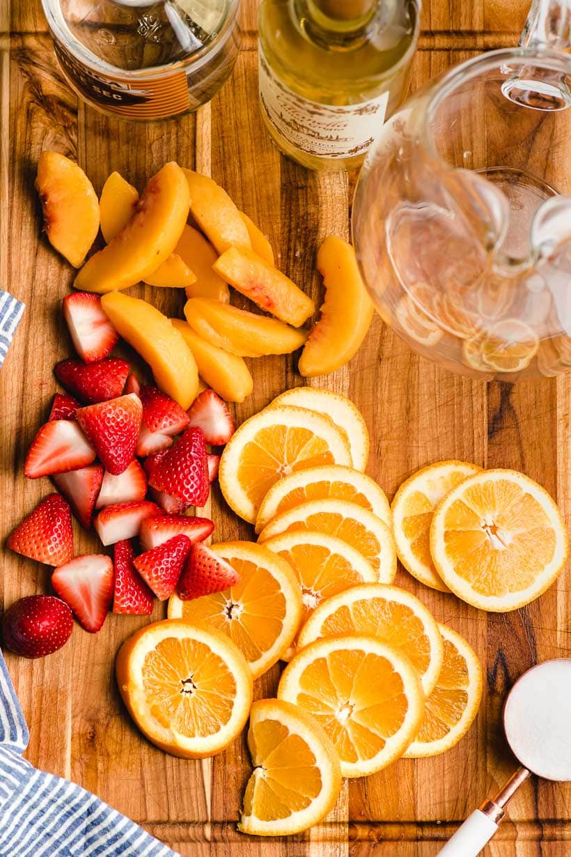 Sliced strawberries, oranges, and peaches arranged on a wood cutting board with orange liqueur and white wine bottles.