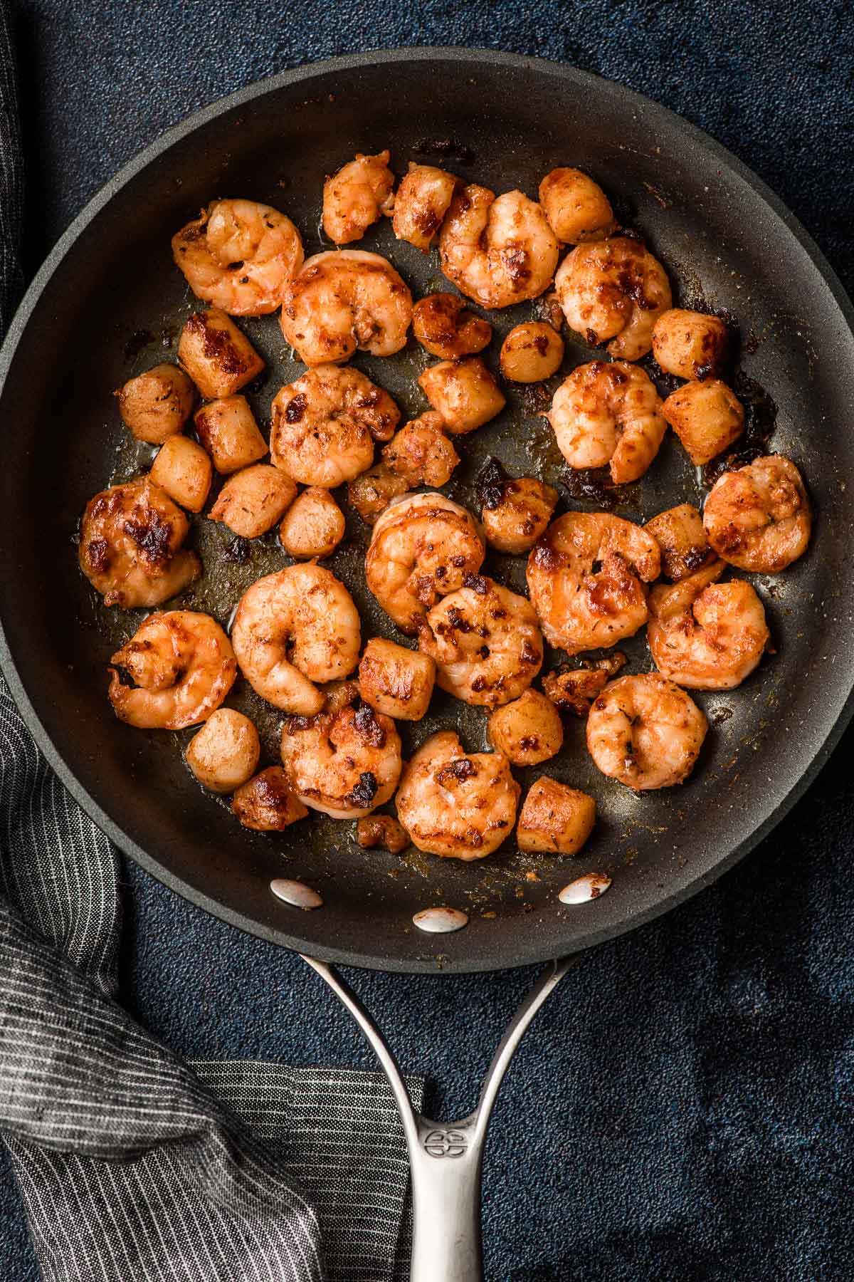 Shrimp and bay scallops shown seasoned and cooked in a dark non stick skillet.