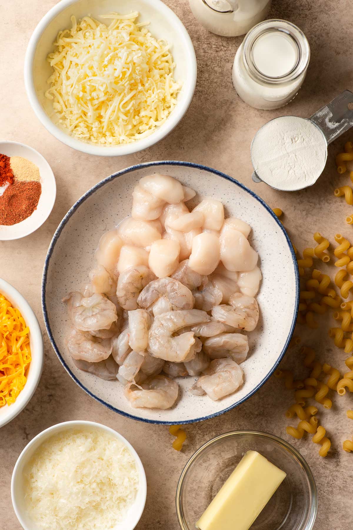 Bowl filled with raw shrimp and scallops, surrounded by other prep bowls with ingredients for mac and cheese-milk, cream, butter, seasonings, and shredded cheeses.