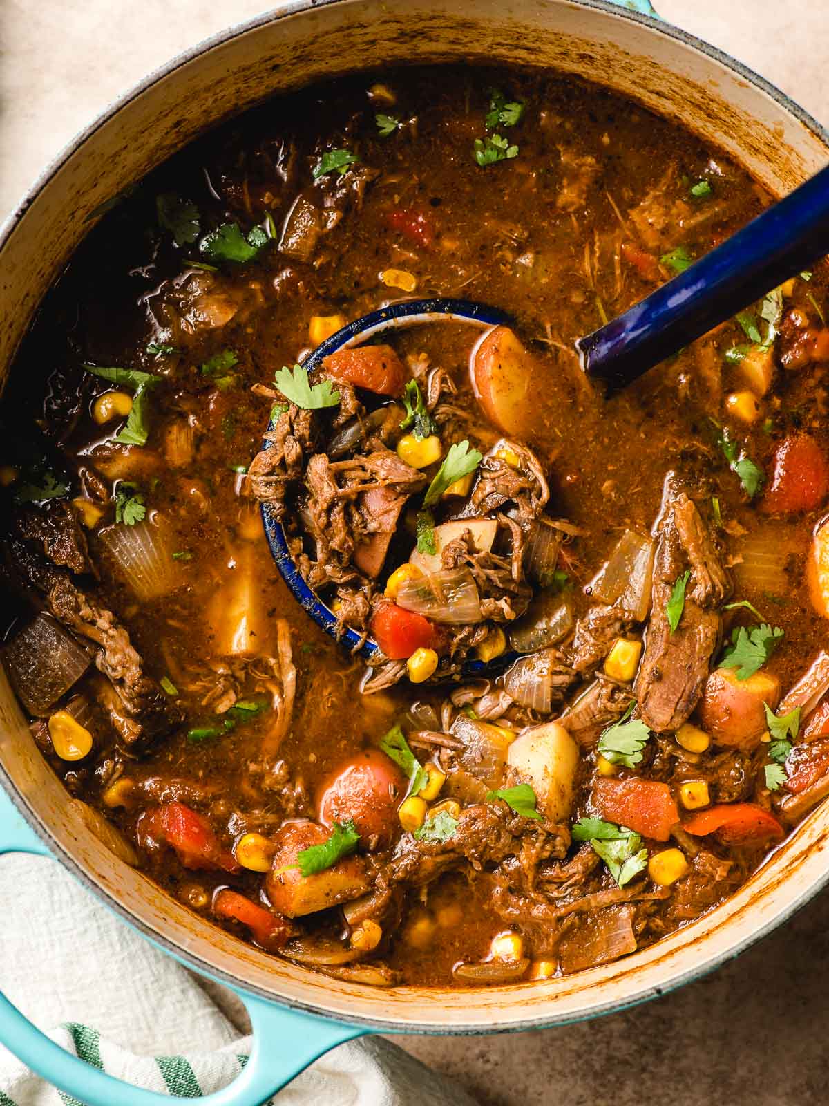 Ladle full of Mexican Beef Stew with barbacoa, potatoes, corn, and tomatoes in a Dutch Oven.